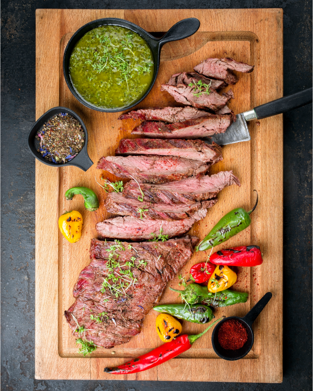 Sliced steak on a wooden cutting board with peppers and chimichurri sauce.