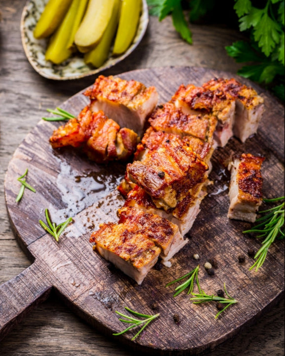 Sliced roasted pork on a wooden board with pickles and herbs on a rustic wooden table.