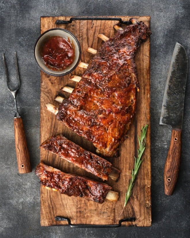 Pork Ribs on a wooden cutting board with a knife and fork on a dark surface