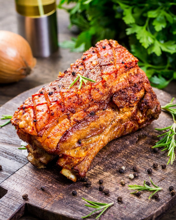 Roasted pork belly on a wooden cutting board with herbs and a bottle of oil in the background.