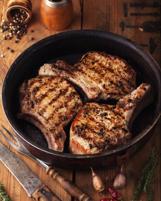 Grilled pork chops in a pan on a wooden table with spices and herbs.